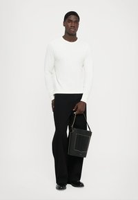Homme portant une chemise blanche à manches longues et un pantalon noir, tenant un sac à main noir avec des détails dorés, se tenant devant un fond blanc uni.