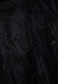 Το μαύρο βελούδινο ύφασμα διαθέτει ένα έντονο λογότυπο «Juicy», διακοσμημένο με λαμπερούς μαύρους στρας, δημιουργώντας μια υφή αντίθεσης στην επιφάνεια.