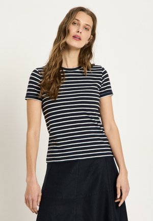 Woman with long wavy hair wearing a black and white striped short-sleeve shirt and a black skirt, standing against a plain background.