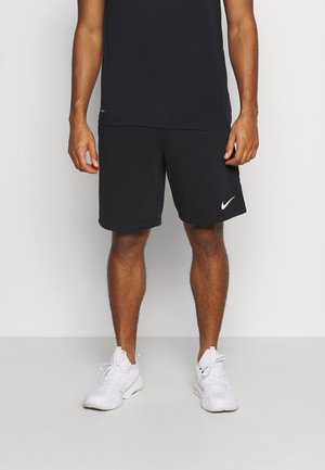 Homme portant un short de sport Nike noir, un t-shirt noir et des baskets blanches, se tenant devant un fond clair.
