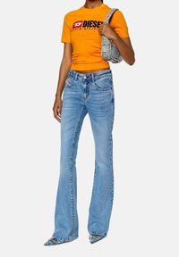 Pomarańczowa dopasowana koszulka z czarnym logo "DIESEL", w połączeniu z jasnoniebieskimi spódniczkami dzwonami i butami na spiczastym czubku. Teksturowana torba przewieszona przez ramię.