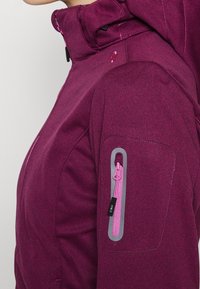 Lila Fleecejacke mit hohem Kragen, einer grauen Reißverschlusstasche am Arm, strukturiertem Stoff und kontrastierendem pinkfarbenem Reißverschlusszug.