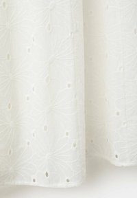 Tissu blanc avec des motifs floraux brodés et de petits œillets, montrant la texture et de légers plis.