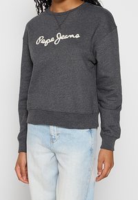 Felpa grigia con collo a girocollo realizzata in misto cotone, con orlo e polsini a coste. Presenta il logo bianco "Pepe Jeans" sul petto. Indossata con jeans azzurri chiari.