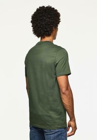 Hombre con pelo rizado que lleva una camiseta de manga corta verde oliva y jeans azules, visto desde atrás ante un fondo blanco liso.