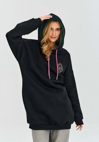 Černá mikina s kapucí lemovanou zelenou barvou, růžovými šnůrkami a grafickým potiskem na hrudi. Materiál se zdá být měkký a pohodlný.