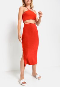 Rotes geripptes Zweiteiler-Set mit einem kurzen Oberteil und einem knielangen Rock mit seitlichem Schlitz. Kombiniert mit weißen Slidesandalen.
