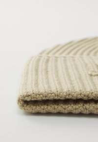 Beige strikket lue med ribbestruktur, har en brettet kant og et mykt, ullaktig utseende. Enkel design uten synlige detaljer.
