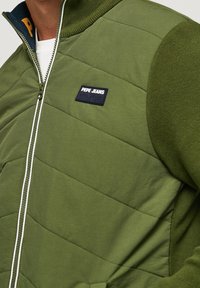 Grön vadderad jacka med dragkedja, med en texturerad tyg, kontrasterande stickad krage och en svart logotyp på bröstet.