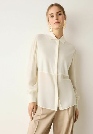 Junge Frau trägt eine transparente weiße Bluse mit Knöpfen und beige Hosen, posiert vor einem schlichten hellen Hintergrund mit goldenen Ohrringen.