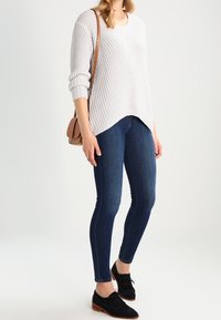 Pull en maille texturée gris clair avec col en V et épaules tombantes, associé à un jean slim bleu foncé et des chaussures noires à lacets. Sac bandoulière marron.