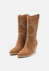 Botas de tobillo de ante marrón con punteras afiladas, costuras decorativas blancas en patrones de sol naciente y tacones de cuña. Textura suave, cremallera en el costado.