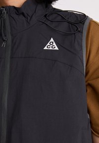 Black sleeveless vest featuring a zip front, ribbed texture, and gray accents. Includes an embroidered logo triangle and adjustable drawstrings.
