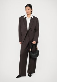 Καφέ υπερμεγέθη σακάκι και ταιριαστά παντελόνια με φαρδιά πόδια πάνω από λευκό πουκάμισο. Μαύρη τσάντα με χρυσές λεπτομέρειες και μαύρες γόβες.