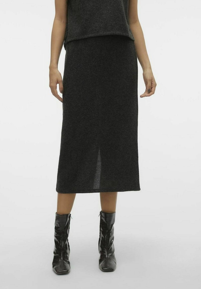 Femme portant une jupe midi en maille gris foncé avec une fente au dos, associée à des bottines noires brillantes sur un fond clair uni.