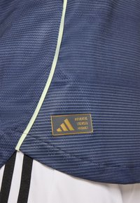 Marinblå idrottströja med texturmönster, accentuerad med ljusgröna detaljer och en Adidas licensierad produktetikett med en gold logotyp.