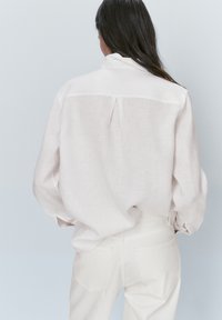 Donna con lunghi capelli scuri che indossa una camicia di lino beige chiaro, morbida e infilata nei pantaloni bianchi a vita alta, in piedi contro uno sfondo chiaro e semplice.