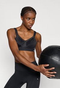 Melns sporta krūšturis ar V veida izgriezumu, acs auduma akcentiem un bieziem siksniņām. Modele tur melnu medicīnas bumbu un valkā melnas legingas.