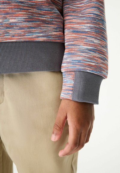 Sudadera de punto multicolor con una superficie texturizada, con puños acanalados grises. Combinada con pantalones beige, mostrando un ajuste relajado.
