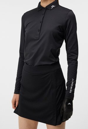 Kobieta ubrana w czarną bluzkę polo z długim rękawem i spódnicę, na prawej ręce czarna rękawica z naszywką "J.LINDEBERG".