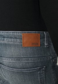 Detailní snímek zadní kapsy modrých denimových džín s hnědou koženou nášivkou s nápisem "DRYKORN" nošených s černým topem.