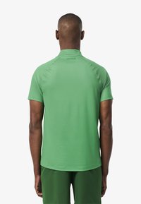 Grön kortärmad polo med rundad krage. Slät textur, figursydd design. "LACOSTE"-logotyp ovanför nacken på baksidan.