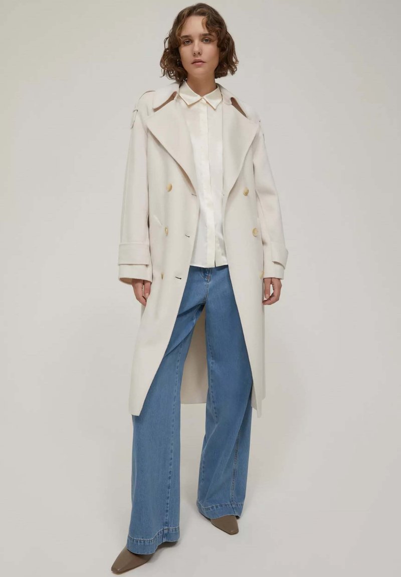 Cappotto lungo color crema con bottoni doppi, indossato sopra una camicia bianca e jeans a gamba larga blu. Tessuto liscio e design su misura.