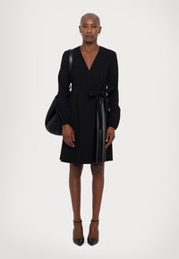 Schwarzes Wickelkleid mit langen Ärmeln, ausgestattet mit einer Seitenschnürung und einem eleganten Satinpanel. Kombiniert mit einer strukturierten schwarzen Handtasche und spitzen Absätzen.