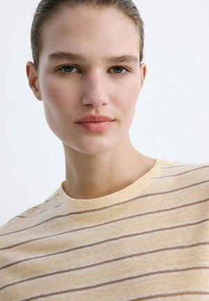 Junge Frau mit heller Haut und blauen Augen, die ein beige gestreiftes Hemd trägt und direkt in die Kamera vor einem weißen Hintergrund blickt.