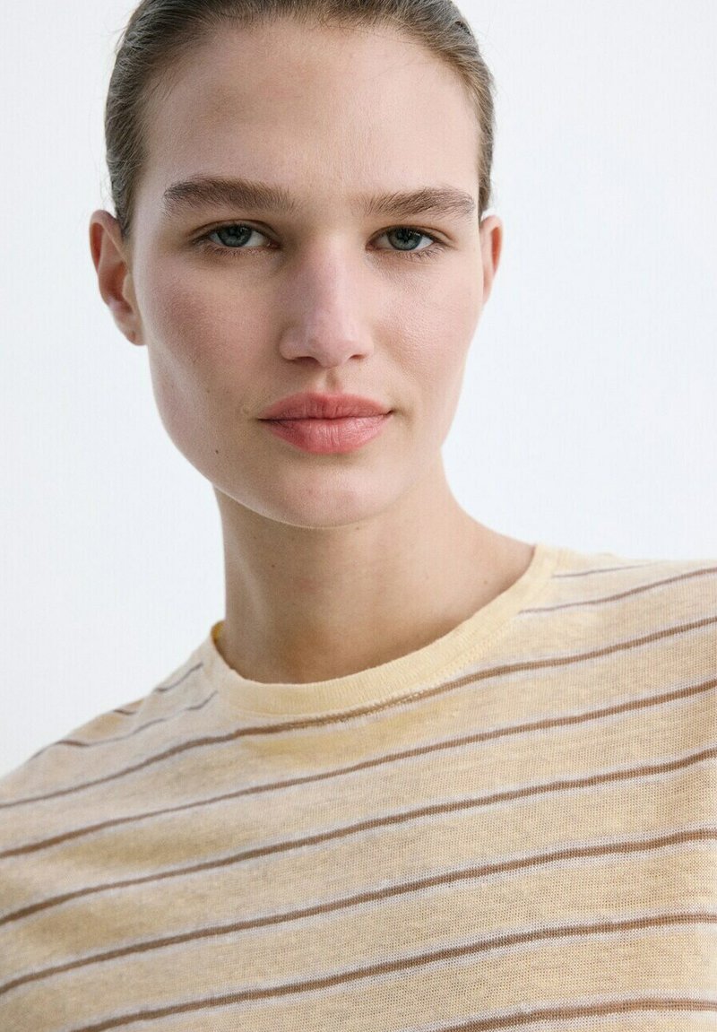 Jeune femme à la peau claire et aux yeux bleus portant une chemise beige à rayures, regardant directement la caméra sur un fond blanc.