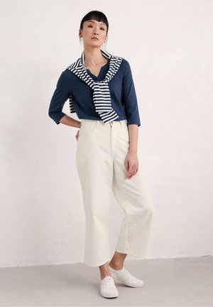 Marineblauwe longsleeve top met een gestreepte stropdas, gecombineerd met crèmekleurige wijde pantalons en witte sneakers op een neutrale achtergrond.