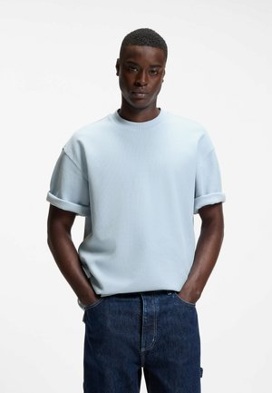 Mężczyzna z krótkimi włosami, ubrany w jasnoniebieską teksturowaną koszulę z krótkim rękawem i ciemnoniebieskie dżinsy, stojący z rękami w kieszeniach.