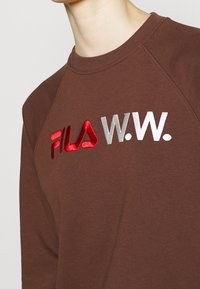 Sudadera marrón con cuello redondo y mangas raglán, adornada con el logo bordado en rojo y plata "FILA W.W." en la parte frontal.