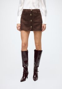 Brown suede mini skirt with front pockets and gold buttons. Paired with glossy, knee-high burgundy boots and a white long-sleeve shirt.