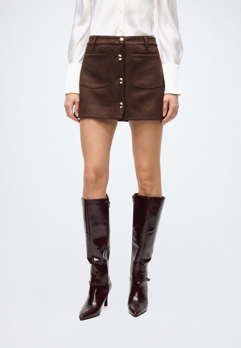 Brown suede mini skirt with front pockets and gold buttons. Paired with glossy, knee-high burgundy boots and a white long-sleeve shirt.