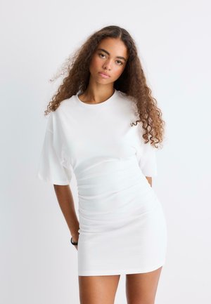 Jeune femme aux longs cheveux bouclés portant une robe blanche ajustée à manches courtes, debout devant un fond clair uni.