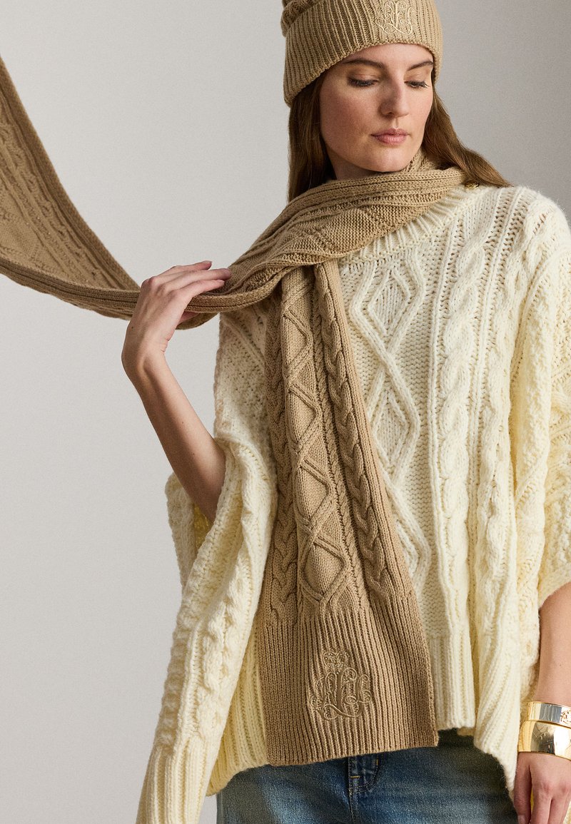 Překládaný krémový oversized svetr s copánkovými vzory, doplněný o texturovanou béžovou šálu a jednobarevnou vroubkovanou čepici s monogramem.