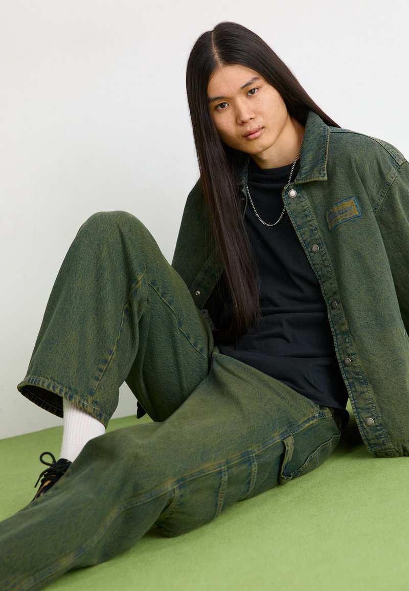 Giacca di jeans verde con una finitura testurizzata, abbinata a pantaloni larghi verdi coordinati. T-shirt nera sotto e calzini bianchi visibili.