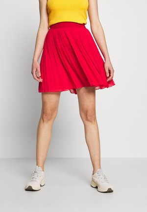 Kobieta w jasnożółtym, bezrękawnym topie, czerwonej plisowanej spódnicy i białych trampkach stoi na jednolitym jasnym tle.