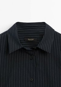Chemise noire à boutons avec fines rayures verticales blanches et col classique, montrant les deux premiers boutons et l’étiquette de marque à l’intérieur du col.
