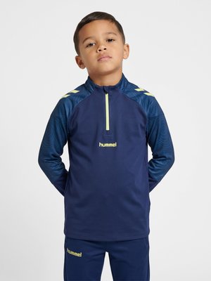 Dreng iført navy blå sportstop og bukser med gul Hummel-logo, stående med hænderne bag ryggen mod en ensfarvet baggrund.