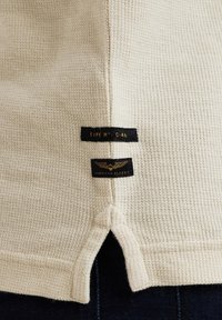 Beige strukturiertes Strickstoff mit gespaltenem Saum, versehen mit schwarzen Etiketten mit goldener Schrift "TYPE N° C-46" und "AMERICAN CLASSIC."