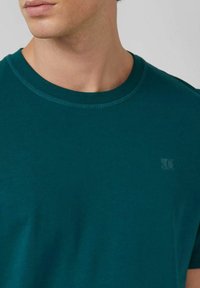 Nahaufnahme einer Person, die ein türkisfarbenes Rundhals-T-Shirt mit einem dezenten Logo auf der linken Brust trägt. Der Hals und die untere Gesichtshälfte sind sichtbar.