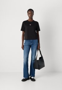 Tricamă neagră scurtă cu un mic logo multicolor, asociată cu blugi evazați de culoare albastră deschisă și o geantă de tote neagră. Material neted, design casual.