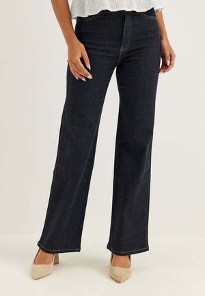 Frau trägt dunkelblaue Jeans mit weitem Bein und beige spitze Absatzschuhe, steht vor einem schlichten hellen Hintergrund.