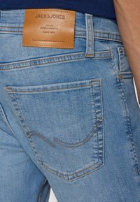 Vaaleansiniset farkut, joissa on nahkainen logoa kuvioitu etiketti, kontrastitikkaukset ja takataskun yksityiskohta.