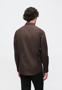 Brun skjorte med lange ærmer, klassisk krave og knapmanchetter, fremstillet i et glat stof og med en buet nederkant for en skræddersyet pasform.