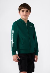 Sudadera verde con cremallera y bolsillo frontal, que presenta el logo blanco de "Champion" en el pecho y la manga. Pantalones cortos negros llevados por debajo.