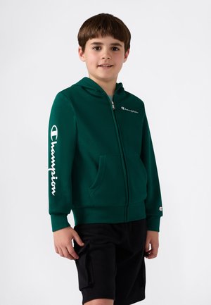 Felpa verde con zip, dotata di tasca frontale, con il logo bianco "Champion" sul petto e sulla manica. Pantaloni corti neri indossati sotto.