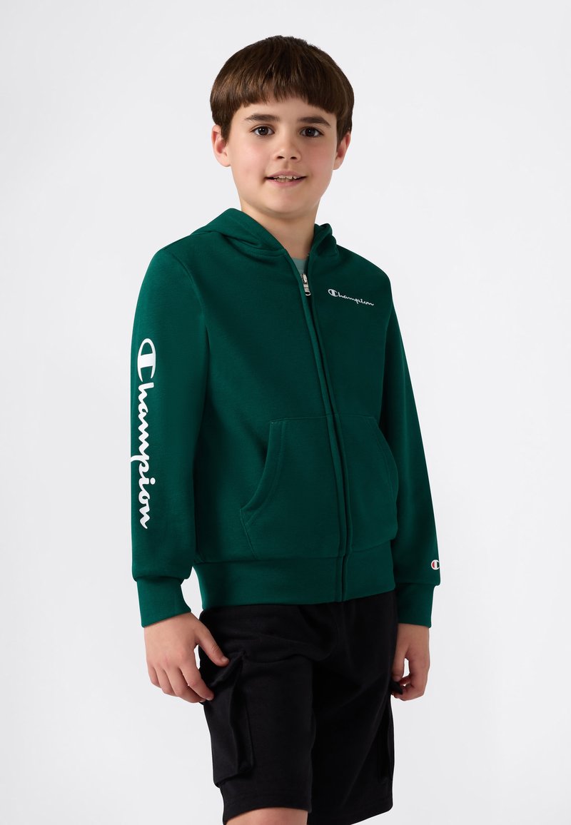 Sudadera verde con cremallera y bolsillo frontal, que presenta el logo blanco de "Champion" en el pecho y la manga. Pantalones cortos negros llevados por debajo.